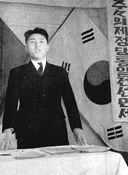 Kim Il-Sung pronunciando un discurso frente a la bandera soviética y el taegukgi en 1946